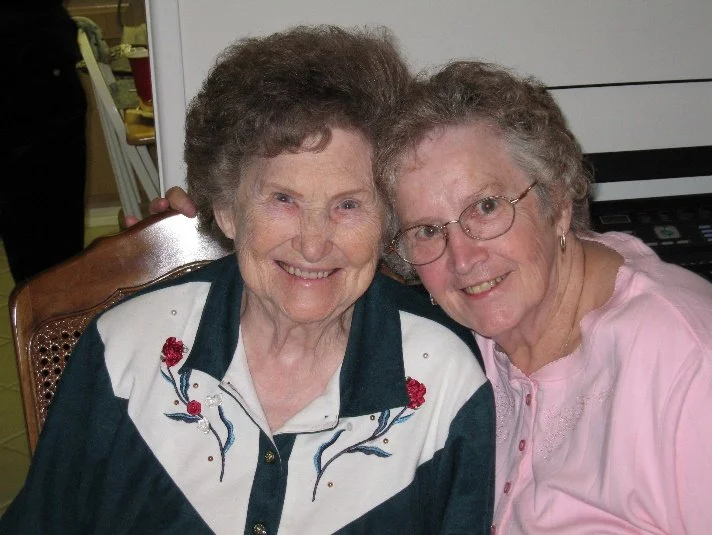 Two elderly women smiling and sitting close together, one wearing a dark green shirt with floral embroidery and the other in a light pink blouse, in an indoor setting.