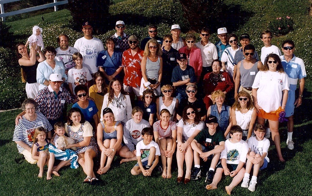 A large group of people, including children, adults, and elderly, gathered outside on a grassy field, posing for a group photo on a sunny day. Some are sitting, others are standing, with a few holding balloons and a woman holding a doll. There are tr