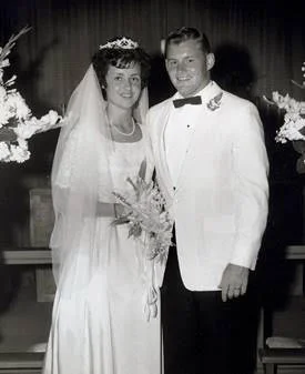 Black and white photo of a bride and groom on their wedding day, standing together indoors with flowers in the background. The bride is wearing a wedding dress and veil, holding a bouquet. The groom is wearing a tuxedo with a bow tie.