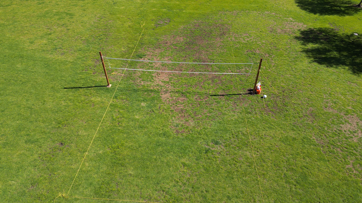 An aerial view of two volleyball poles set up on a grassy field, with a person in an orange shirt sitting near one of the poles.