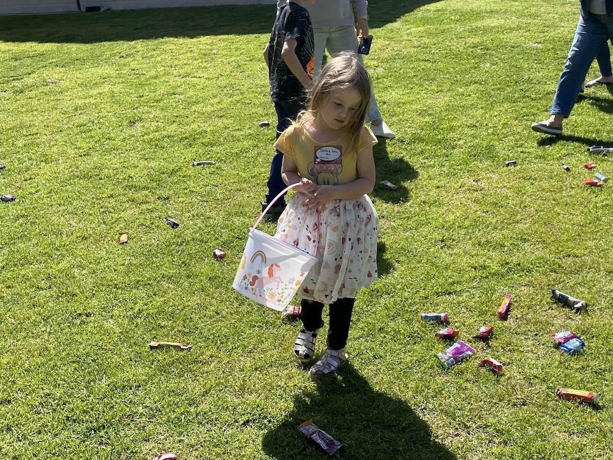 A young girl in a yellow shirt and floral skirt holding a colorful bag walks on a grassy field scattered with candy wrappers. Other children and adults are in the background.