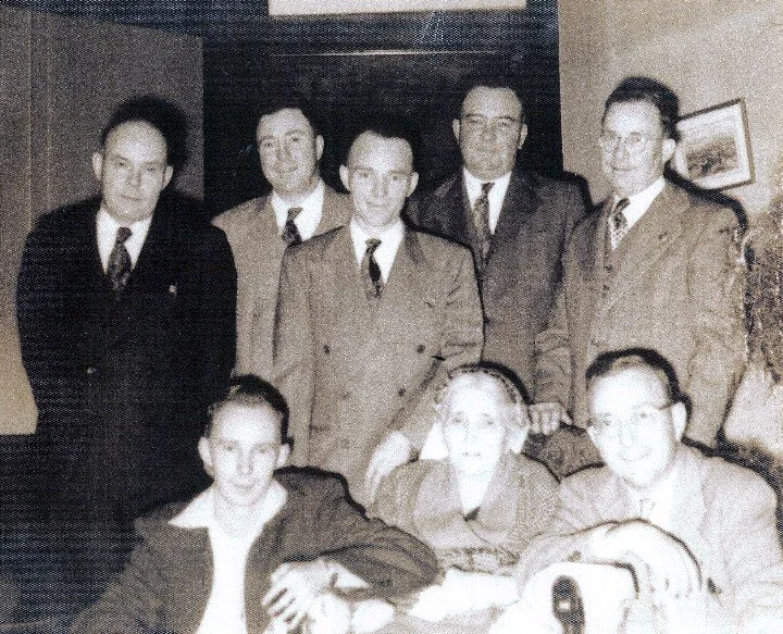 Seven men, five standing and two sitting, in a room with framed pictures on the wall, dressed in suits and military uniforms, from a vintage black-and-white photograph.