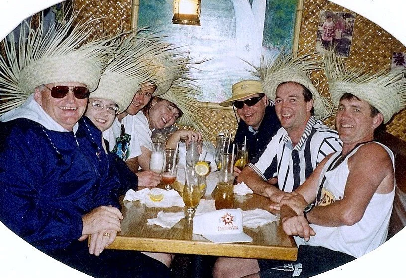 Seven people sitting around a restaurant table, wearing large straw hats and sunglasses, enjoying drinks and smiling.