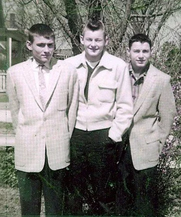 Three young men standing outdoors in front of trees, dressed in suits and blazer jackets, posing for a photo.