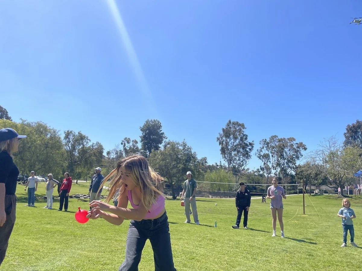 Group of people playing a game with balloons on a sunny day at a park, with trees and blue sky in the background.