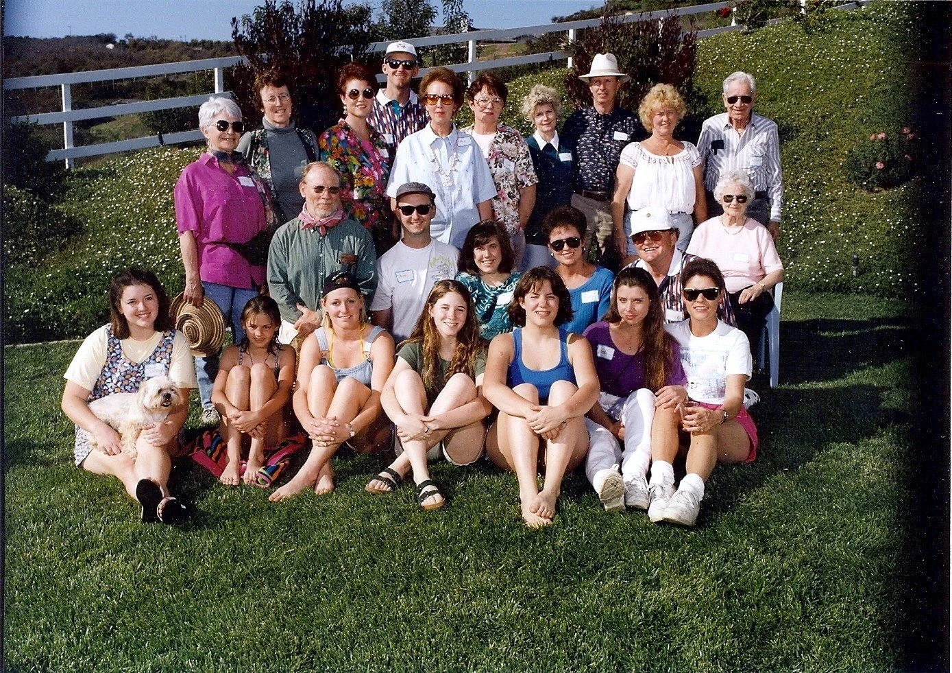 A group of people, including children, young adults, and seniors, gathered outdoors on a grassy yard for a group photo during daytime. Some are sitting on the grass, some are standing in the back, with a few holding hats or wearing sunglasses. There 