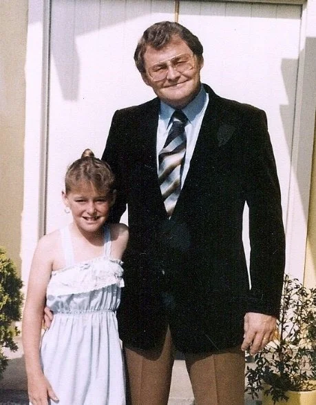 A man in a suit and a girl in a white dress standing together outside in front of a house, smiling.