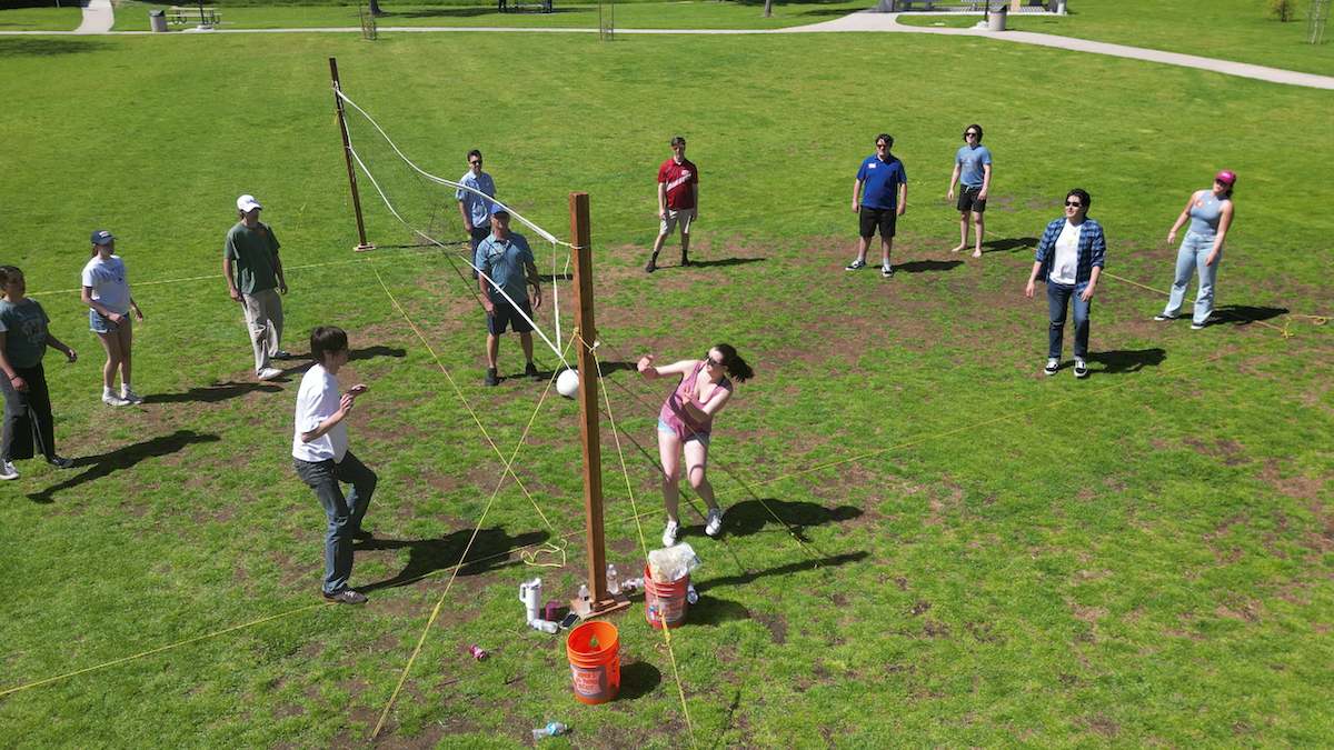 People playing a game of volleyball outdoors on a grass field, with some wearing casual clothing and sunglasses, on a sunny day.