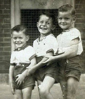Three young children smiling and holding hands outdoors in front of a brick building with a wire window.