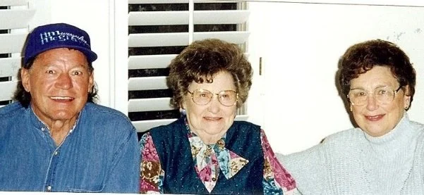 Three older women sitting together indoors, smiling, with a window with blinds in the background.