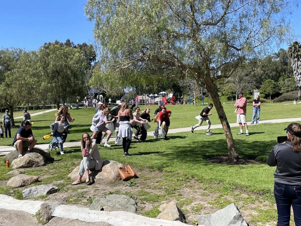 People playing a game of push the penny in a park on a sunny day with trees, grass, and a playground in the background.