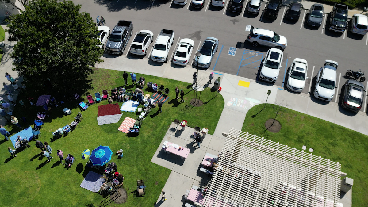 An aerial view of a park with people gathered for a daytime outdoor event. There are several tents, tables, and umbrellas set up on the grassy area, with people sitting, standing, and walking around. Adjacent to the park is a parking lot with numerous parked cars.