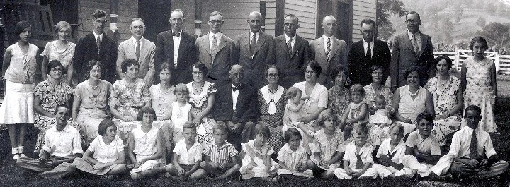 Group photo of children and adults, with boys wearing white shirts and dark pants, girls in dresses, and men in suits, taken outdoors in front of a brick building.