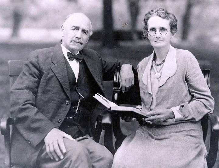 A black-and-white photo of an older man and woman sitting together on a park bench. The man is wearing a suit with a bow tie and the woman is dressed in a long skirt and blazer, holding a book or folder in her lap.