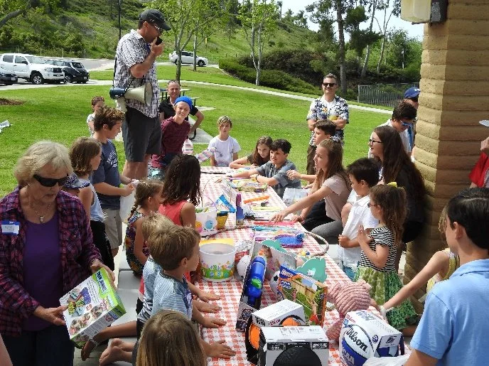 A diverse group of children and adults gathered outdoors around a long table with various items, participating in a community event or celebration.