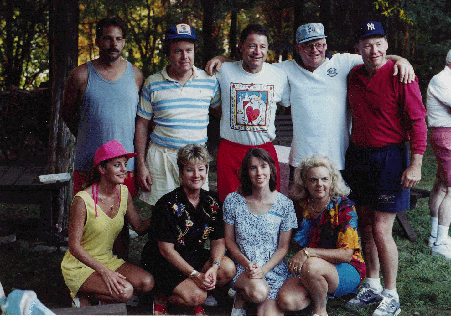 A group of nine people, five men standing in the back row and four women sitting in the front row, posing outdoors in a wooded area during daytime. The group is dressed casually, with some wearing colorful or patterned clothes.