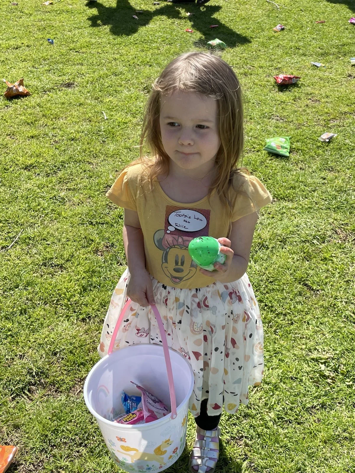A young girl standing outdoors on grass, holding a green plastic egg and a pink and white bucket filled with candy and toys. She is wearing a yellow T-shirt with a Minnie Mouse graphic, a skirt with colorful patterns, and sandals. There are scattered