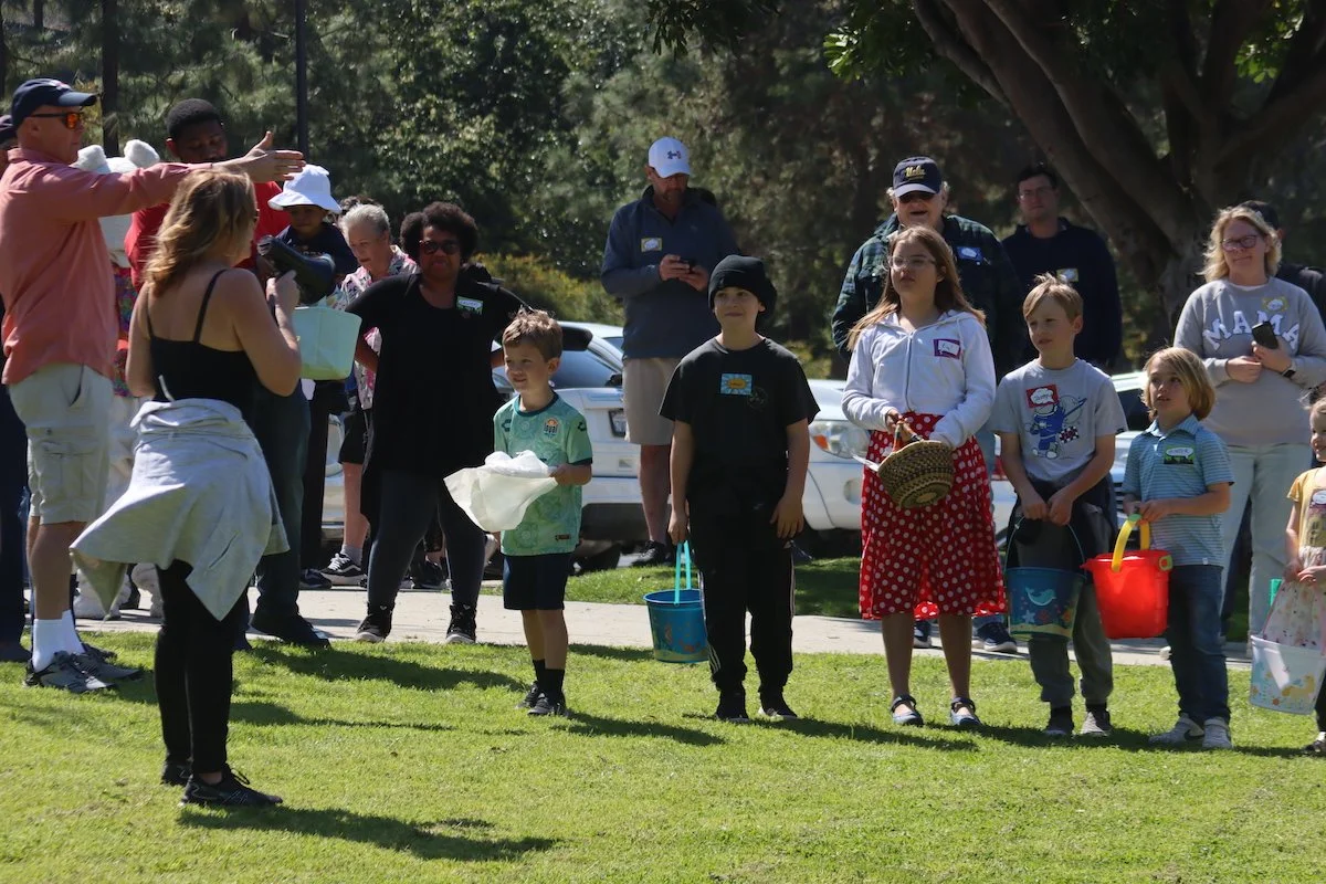 Group of children and adults gathered outdoors for an Easter egg hunt, with children holding baskets and eggs, and adults supervising.