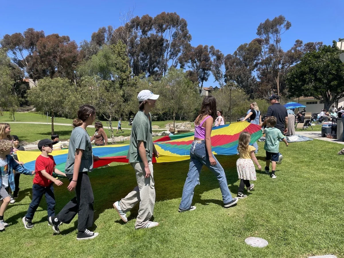 Children and adults playing with a large rainbow-colored parachute in a park on a sunny day.