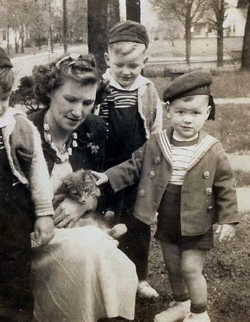 Black and white photo of a woman and two young boys outdoors, with the woman holding a kitten. The boys are wearing caps and jackets, and there are trees and a park in the background.