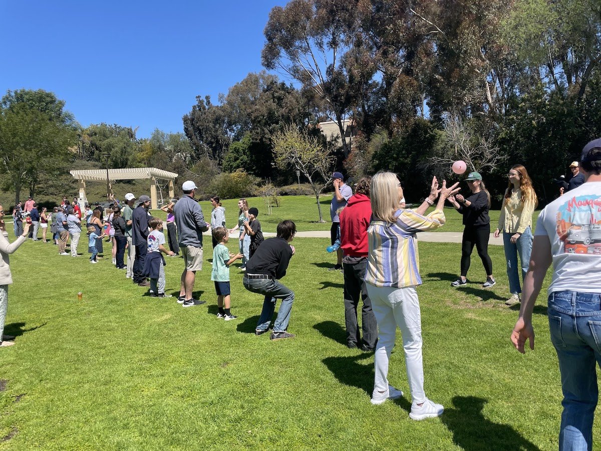 People standing in a line outdoors on a bright, sunny day in a park, some playing with balls and children.