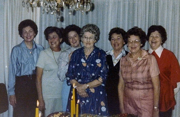 Group of seven women celebrating an occasion with a cake and candles, standing indoors in front of a curtain and chandelier.