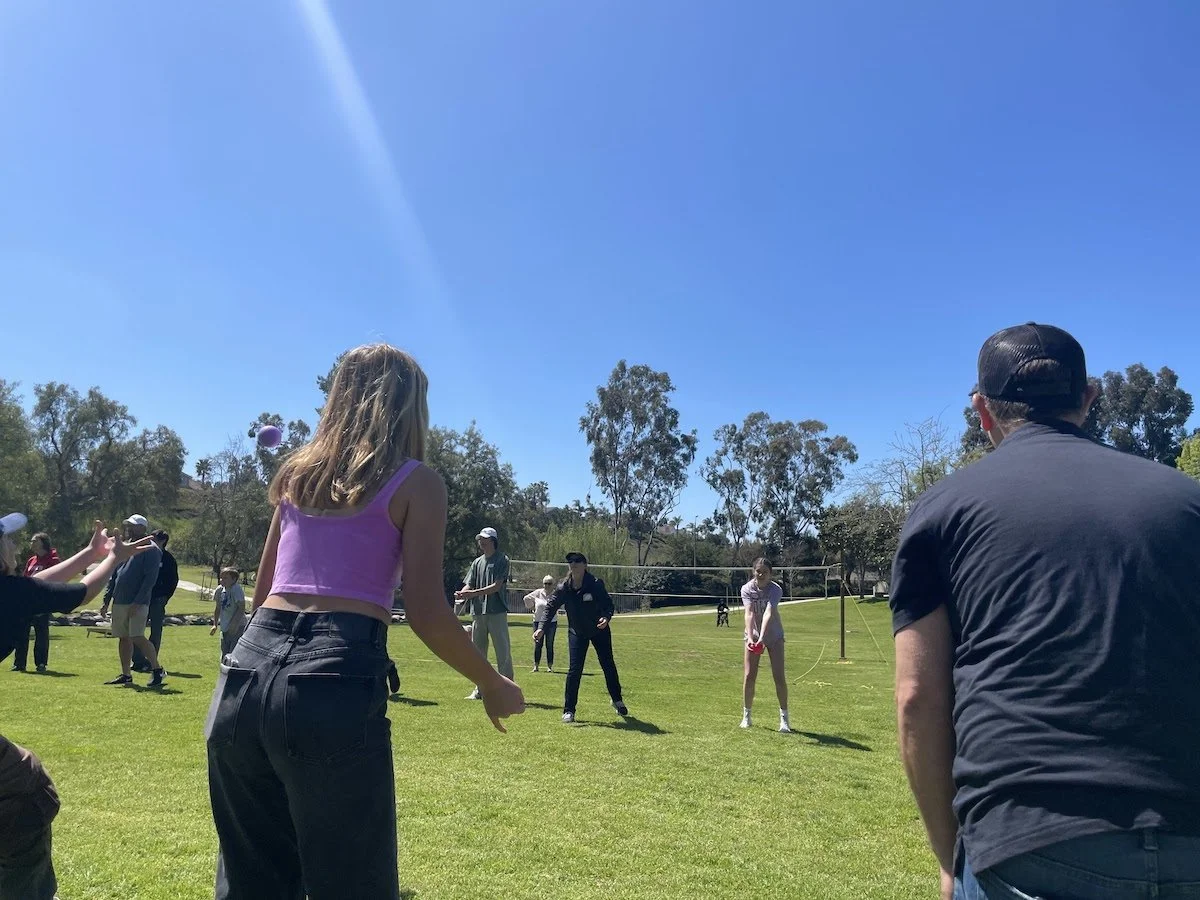 People playing volleyball outdoors on a sunny day in a grassy field with trees in the background.
