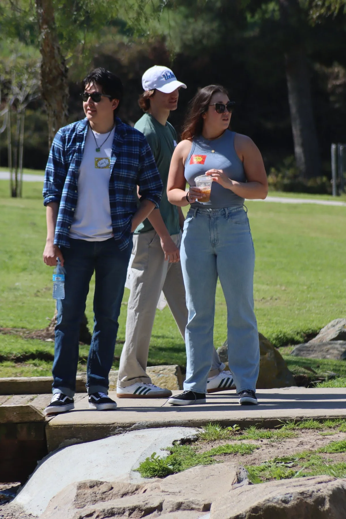 Three young adults standing outdoors on a sunny day in a park-like setting with trees and grass, casually dressed and wearing sunglasses.