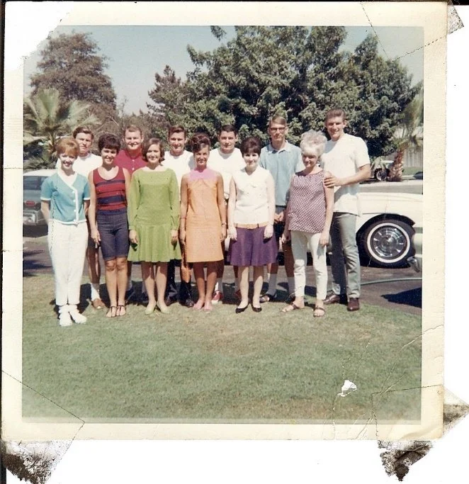 Group of twelve people standing outdoors on a lawn, with cars and trees in the background, posing for a photo in vintage clothing.