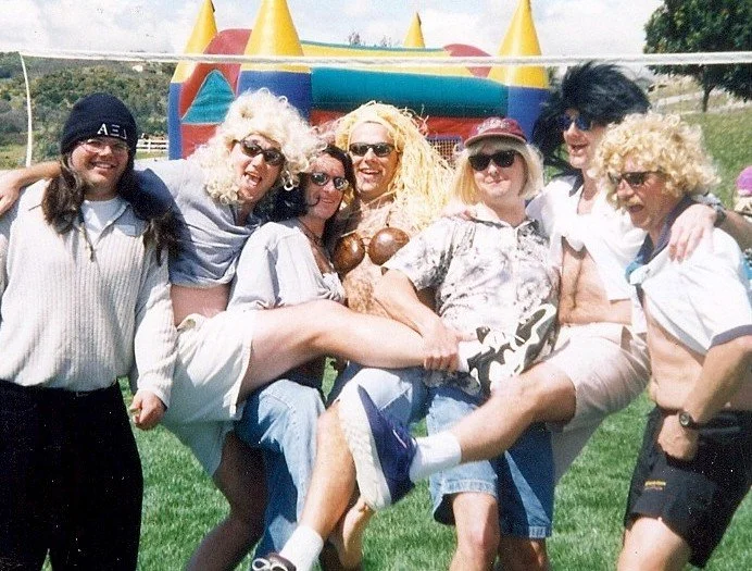 Group of seven women outdoors standing on grass, smiling, some wearing wigs, sunglasses, and vintage-style clothing, with inflatable bounce house in the background.