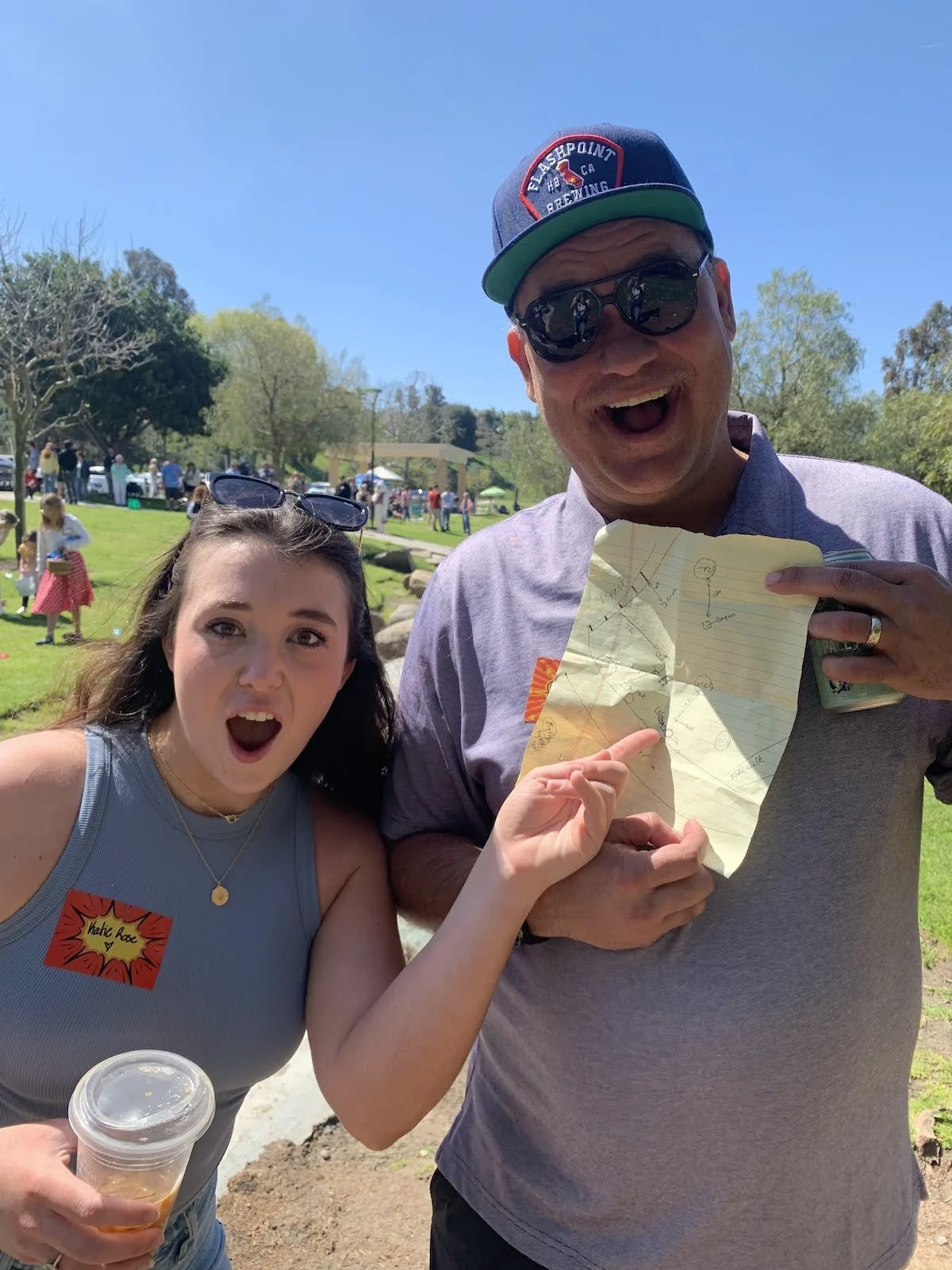 Two people at an outdoor event, a woman and a man, holding a crumpled piece of paper with diagrams, with trees and other people in the background on a sunny day.