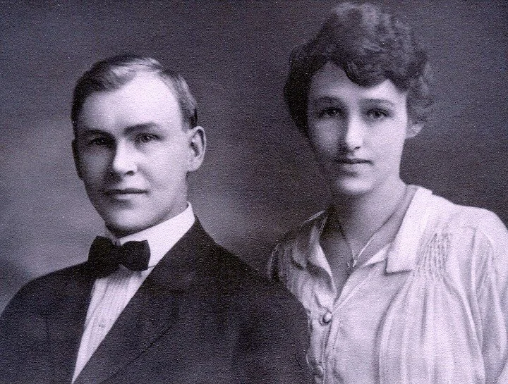 A black and white portrait of a man in a tuxedo with a bow tie and a woman in a light-colored blouse with a necklace, both looking at the camera.