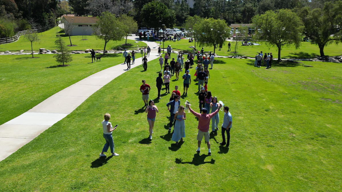 Group of people gathered on a grassy park with a curved sidewalk, trees, and a parking lot in the background. Some are standing in a circle, others are walking around.