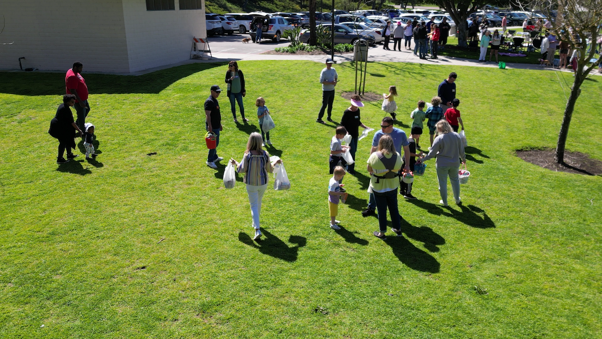 People gathering outdoors on a grassy area, some holding bags and children, with a parking lot and trees in the background.