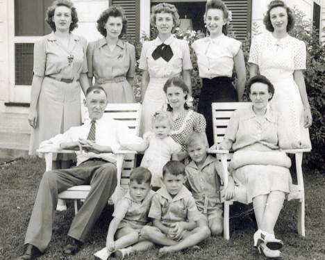 A black-and-white photo of ten people outdoors, including a man and a woman seated in chairs with children around them, and six women standing behind them, in front of a house with windows and shutters.