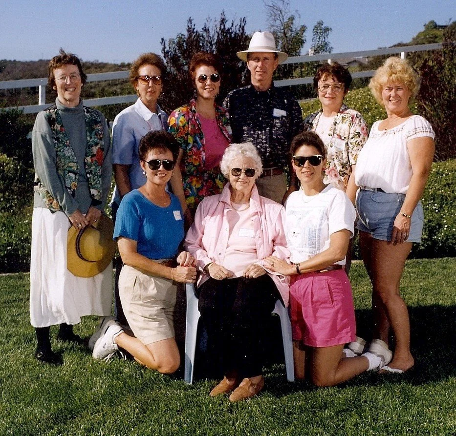 Group of ten women and one man posing outdoors on a lawn with greenery and hills in the background, all smiling.