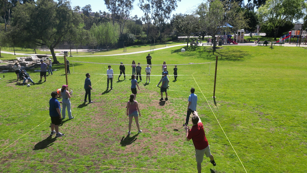 People playing volleyball on a grassy park field with playground equipment and trees in the background.