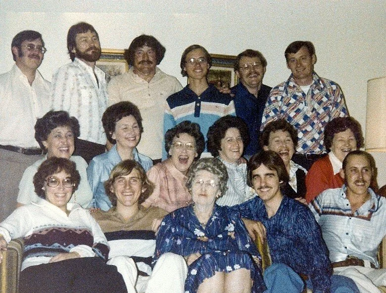 Group of people gathered indoors for a photo, including women and men of various ages, some seated and some standing behind, smiling participate in a casual social setting.