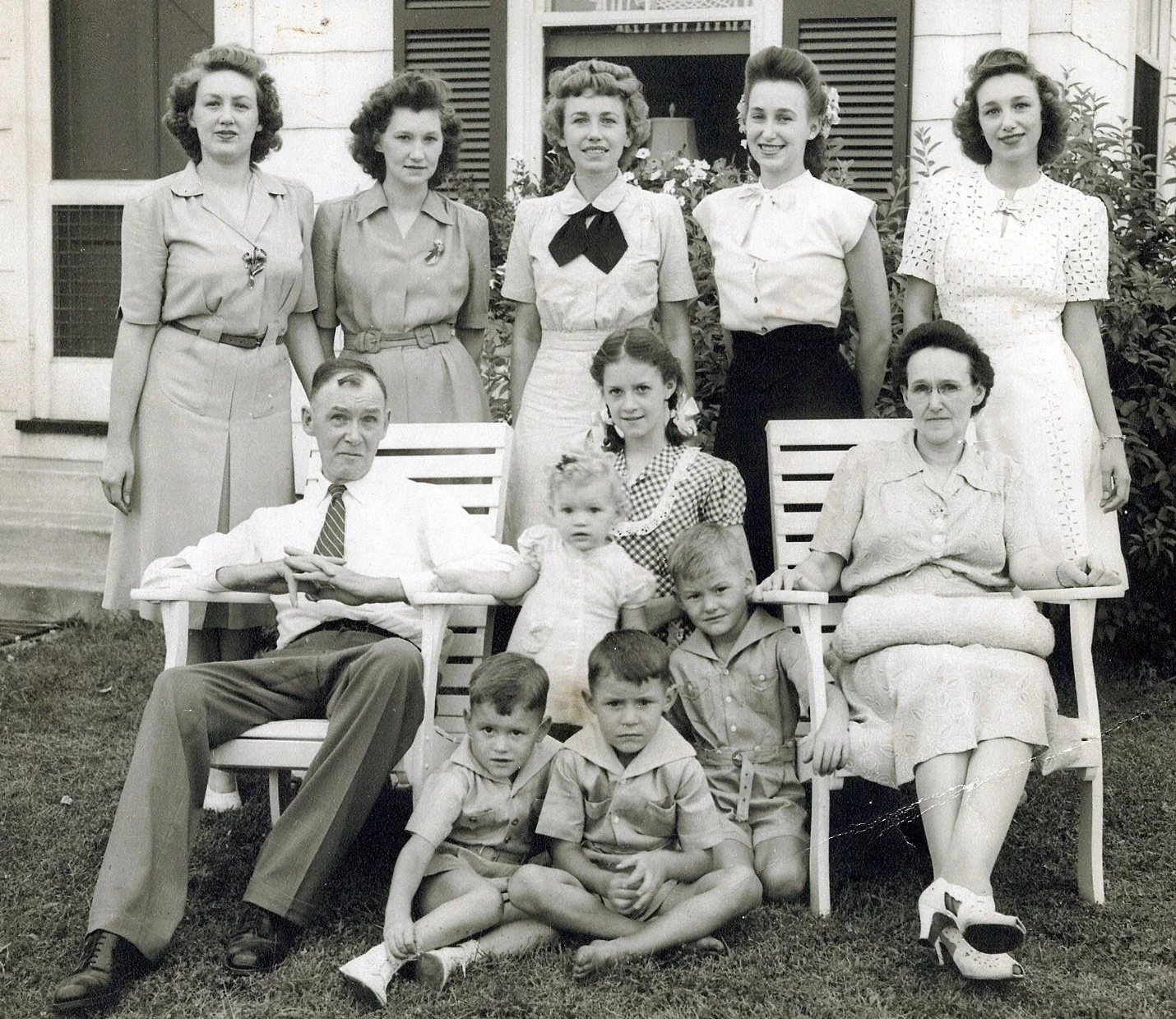 A black-and-white photograph of a family group outside a house. There are three adults sitting on chairs, surrounded by seven children and five women standing behind them. The setting appears to be in front of a house with a door and window with open
