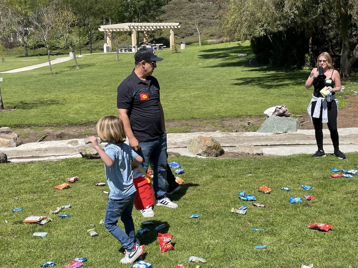 A woman taking a photo with a camera, a man and a young boy standing on a grassy park area with scattered snack bags, trees, a pavilion, and walking paths in the background.