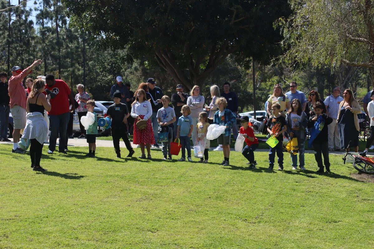 Children and adults standing in line outdoors on a grassy field, participating in an event with some holding containers and toys, under trees with more people and parked cars in the background.