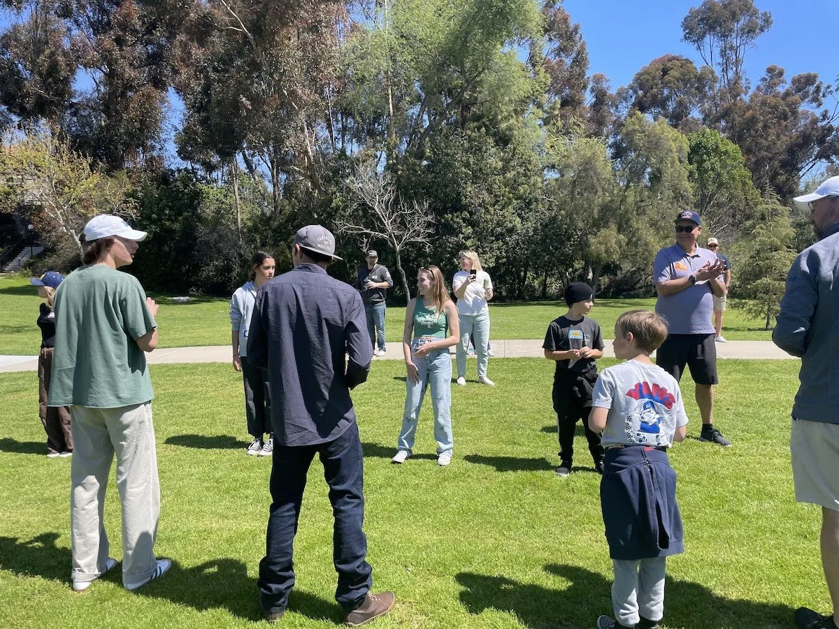 Group of people, including children and adults, gathered outdoors on a sunny day in a park with green grass and trees.