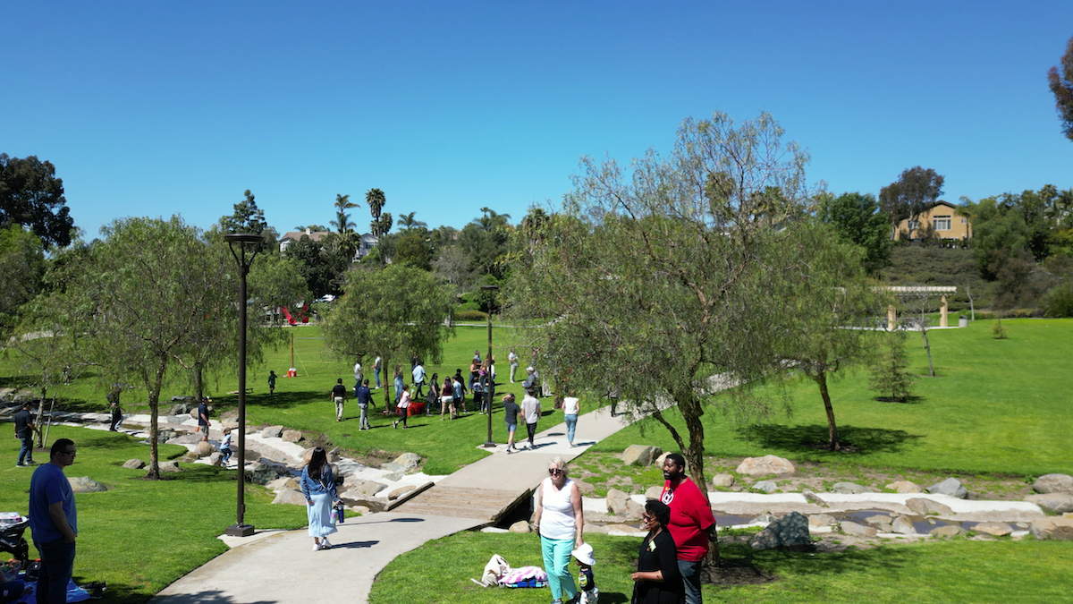 People gathering in a park with green grass, trees, a small stream, and a clear blue sky.