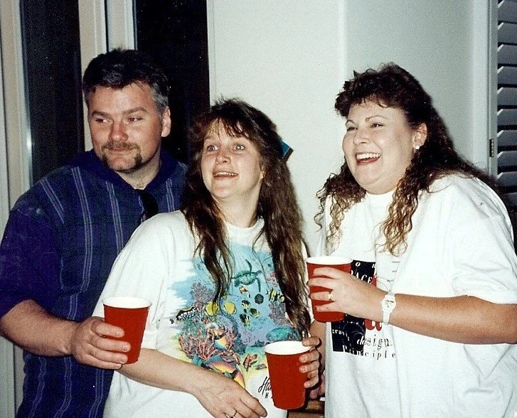 Three people, two women and one man, smiling and holding red cups, standing indoors near a window.