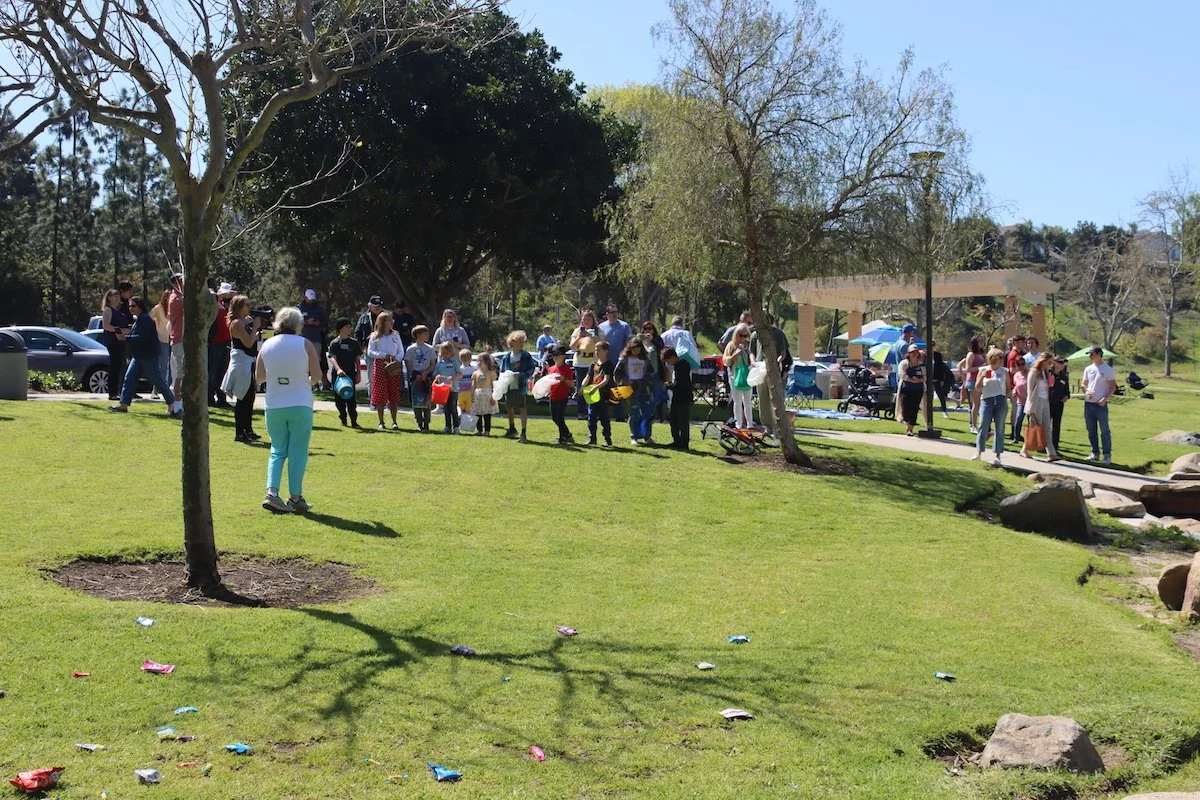 People in a park lined up for an event, with children holding containers and adults supervising under trees and near a pavilion.