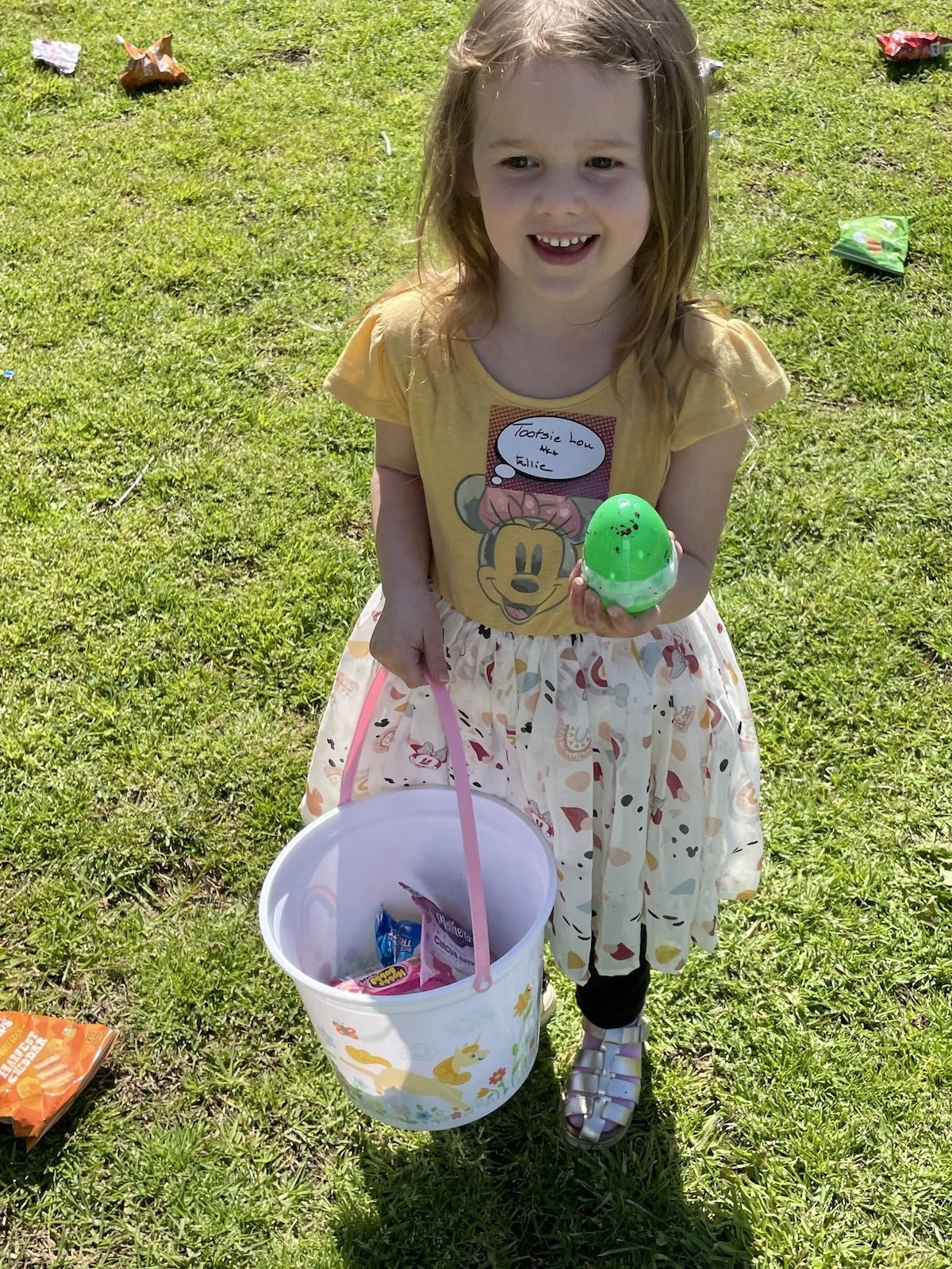 A young girl with light brown hair smiling outdoors on a grassy field. She is wearing a yellow T-shirt with Minnie Mouse on it and a colorful skirt. She is holding a green plastic egg in one hand and a pink basket with candy in the other. There are c