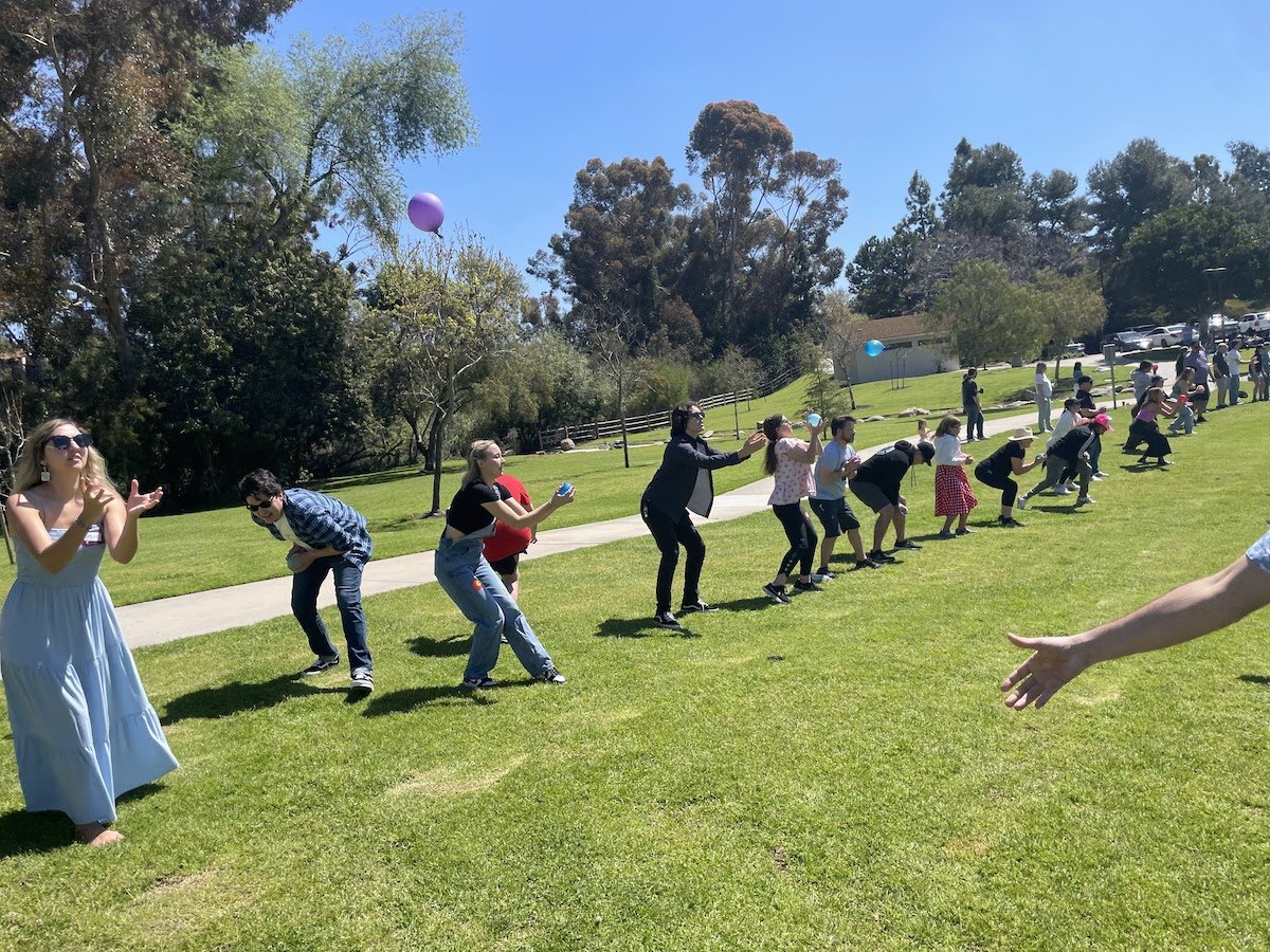 People outdoors on a sunny day participating in a game of catch with balloons, some reaching to catch the balloons in the air, on a grassy field with trees and parking lot in the background.