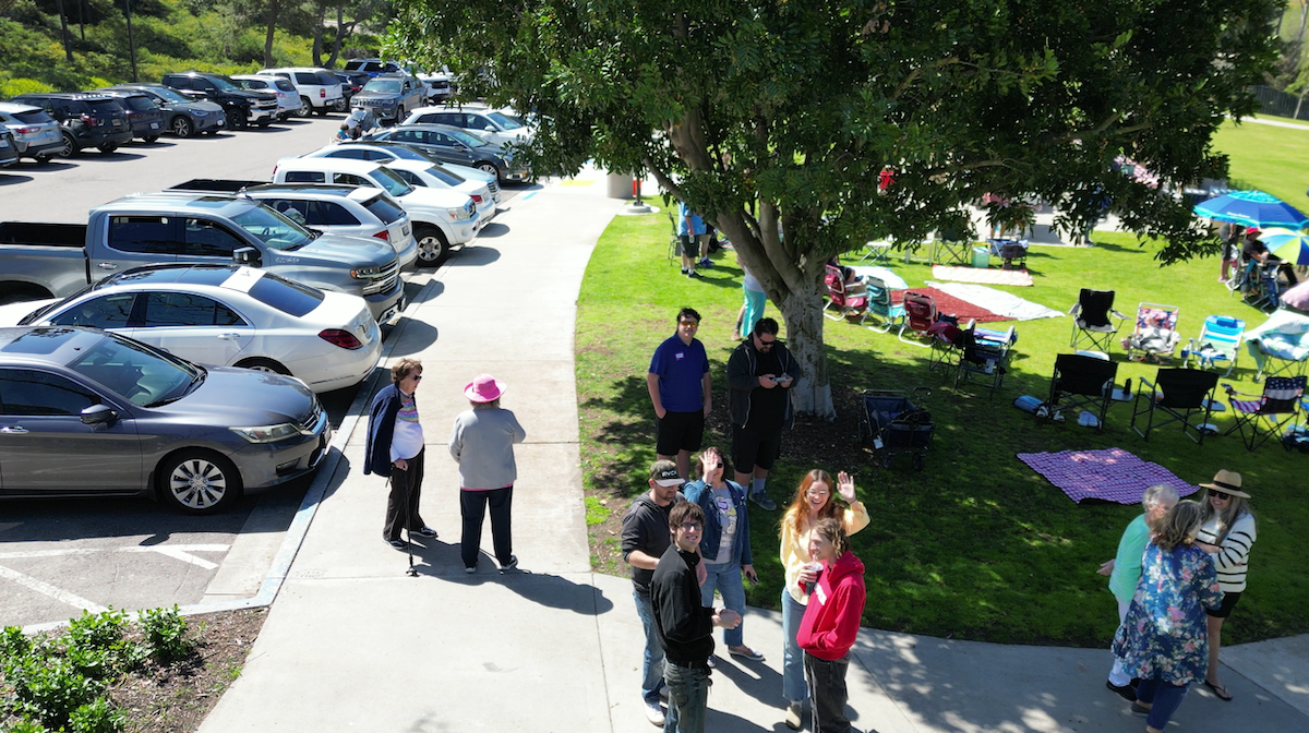 People gathering in a park with grassy areas, picnic blankets, and chairs. Several cars are parked along a driveway, and trees provide shade.