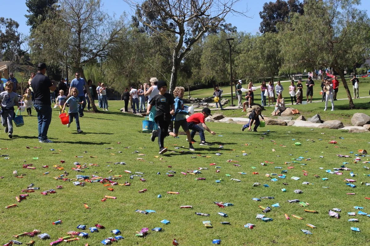 People participating in an Easter egg hunt in a park, with children collecting plastic eggs scattered across the grass.