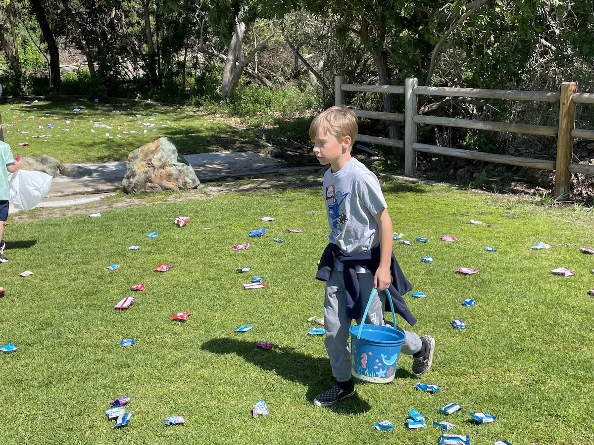A boy holding a blue bucket with a sea creature design walks across a grassy area scattered with candy wrappers on a sunny day. Another child is partly visible on the left side of the image.